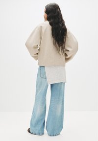 Giacca beige oversize con spalle scese sopra un maglione grigio chiaro a maglia, abbinata a jeans larghi blu chiaro con una finitura sbiadita.
