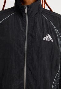 Veste de sport noire à col haut, fermeture zippée, accents argentés, tissu texturé et logo brodé sur la poitrine gauche.