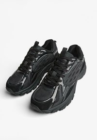 Čierne športové topánky so sieťovinou a syntetickými materiálmi, polstrovaným golierom a šnurovaním. Výrazné textúrované akcenty a robustná podrážka.