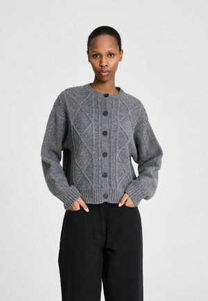Personne debout de face, portant un cardigan gris boutonné avec un motif losange et un pantalon noir, mains dans les poches, fond neutre.