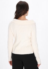 Crème-kleurige kabelgebreide trui met een brede, geribbelde off-the-shoulder kraag en lange mouwen. De textuur is zacht en dik.