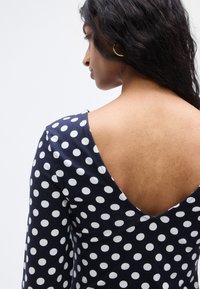 Μπλε σκούρο μπλούζα μακρυμάνικη με λευκές πουά, με βαθύ V στο πίσω μέρος και λεία υφή. Ορατό χρυσό σκουλαρίκι σε σχήμα κρίκου.