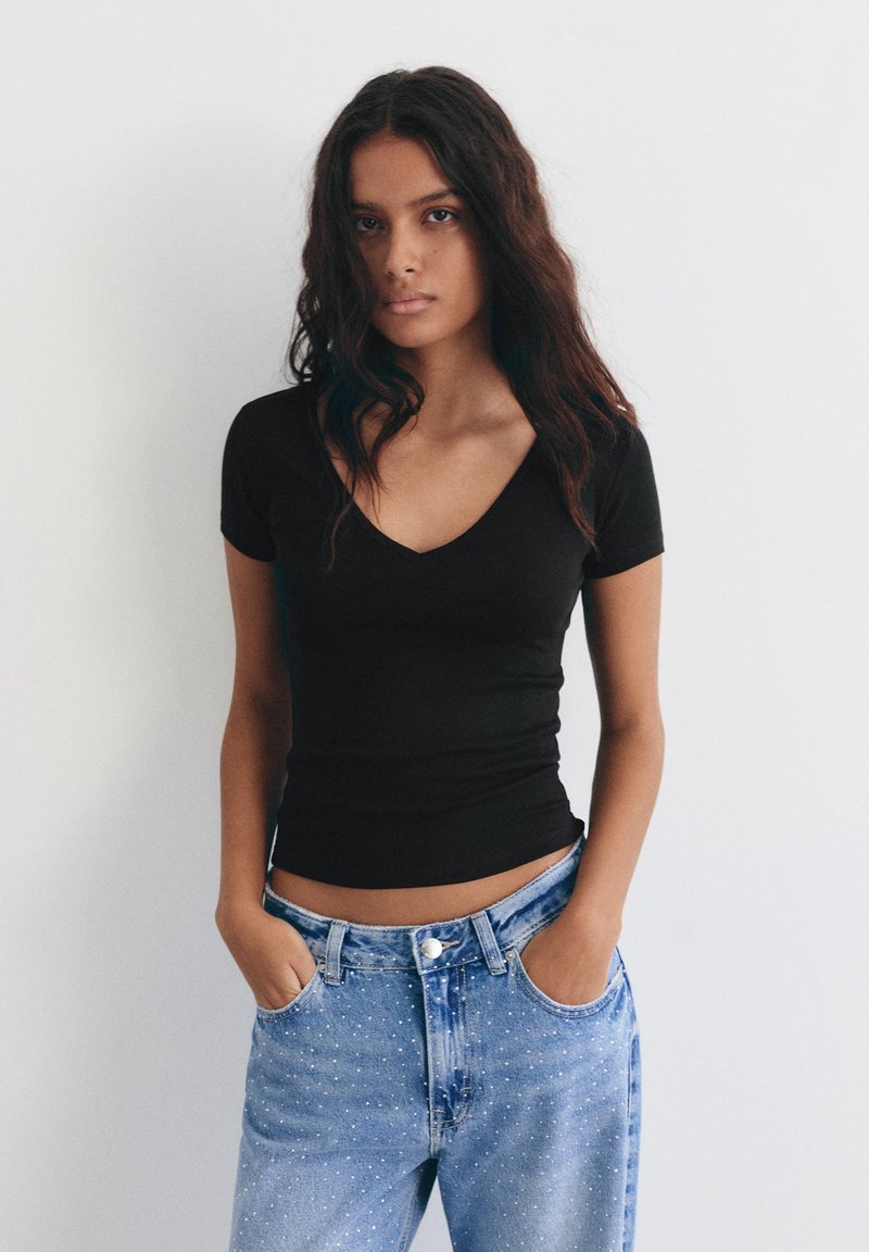 Jeune femme aux cheveux longs et foncés portant un haut noir à manches courtes et encolure en V, et un jean bleu clair, les mains dans les poches, debout devant un fond uni.