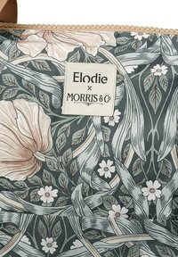 Stof med blomsterprint, der har store ferskenfarvede blomster og grønne blade. Inkluderer et beige label med "Elodie x MORRIS & CO."