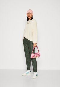Creme strikket sweater, olivengrønne bukser, lyserød strikket hue og en lyserød mønstret taske med guldhardware, båret med hvide sneakers.