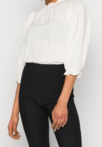 Blouse blanche avec des accents de volants plissés, manches bouffantes longues et décolleté rond, associée à un pantalon noir taille haute.