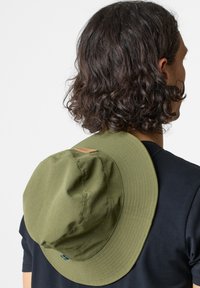 Cappello a secchiola verde oliva con un'ampia visiera, realizzato in tessuto morbido. Include un piccolo etichetta in pelle e dettagli cuciti, indossato all'indietro.