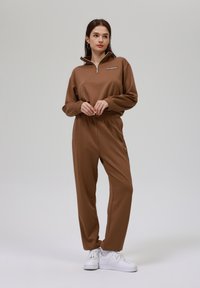 Rjav zip-up pulover s elastičnimi manšetami in pasom, v kombinaciji z usklajenimi ozkimi hlačami; ima mehak teksturo in bele superge.
