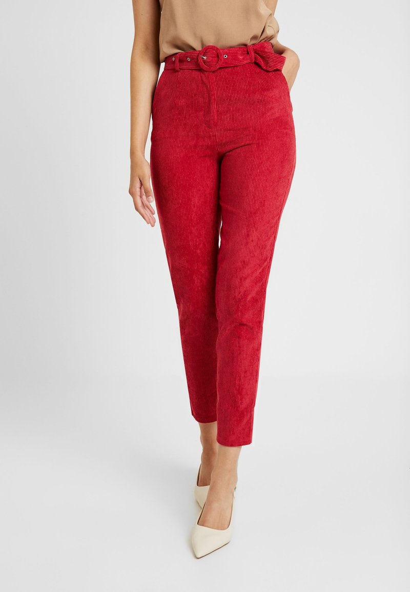 Femme portant un pantalon en velours côtelé rouge taille haute avec ceinture assortie et des talons beiges à bout pointu, debout devant un fond clair uni.