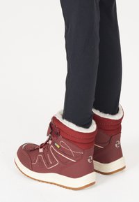 Bottes d'hiver rouges texturées avec garniture en fausse fourrure, fermeture à lacets et semelle en caoutchouc. Assorties à des leggings noirs avec des coutures verticales.
