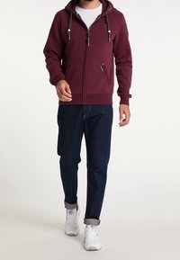 Sudadera burdeos con cremallera, cordones, dos bolsillos frontales, puños acanalados y una capa interior blanca, combinada con jeans oscuros y zapatillas blancas.