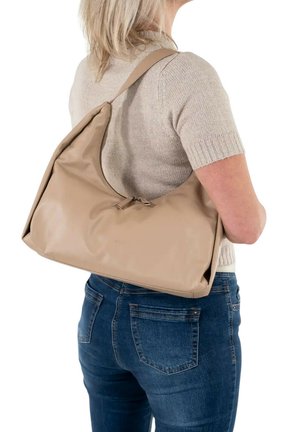 Persona bionda con maglione beige a maniche corte e jeans blu porta una grande borsa beige con cerniera a tracolla sulla schiena.