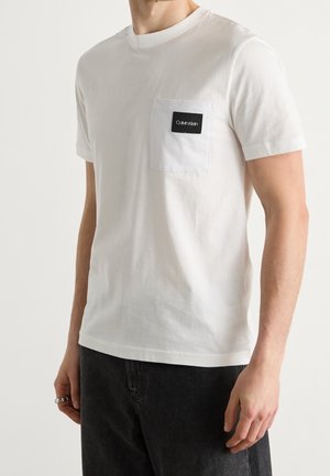 Mann trägt ein weißes kurzärmeliges T-Shirt mit einer Brusttasche, die mit einem schwarzen Calvin Klein Logo-Patch versehen ist.
