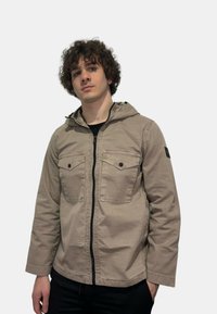 Giacca marrone chiaro con cappuccio, dotata di due tasche sul petto con pattina, zip frontale e vestibilità comoda. Realizzata in tessuto testurizzato.
