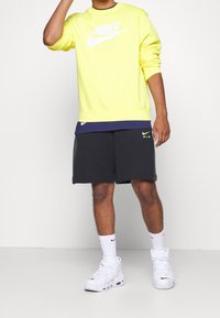 Gul sweatshirt med vit Nike-logotyp, över en marinblå skjorta. Svarta shorts med grön Nike Air-logotyp, tillsammans med vita sneakers.