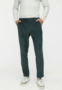 Férfi sötétzöld kordbársony nadrágban, fehér hosszú ujjú pólóban és szürke díszítéssel ellátott fehér sportcipőben, zsebeiben tartja a kezét, fehér háttér előtt áll.