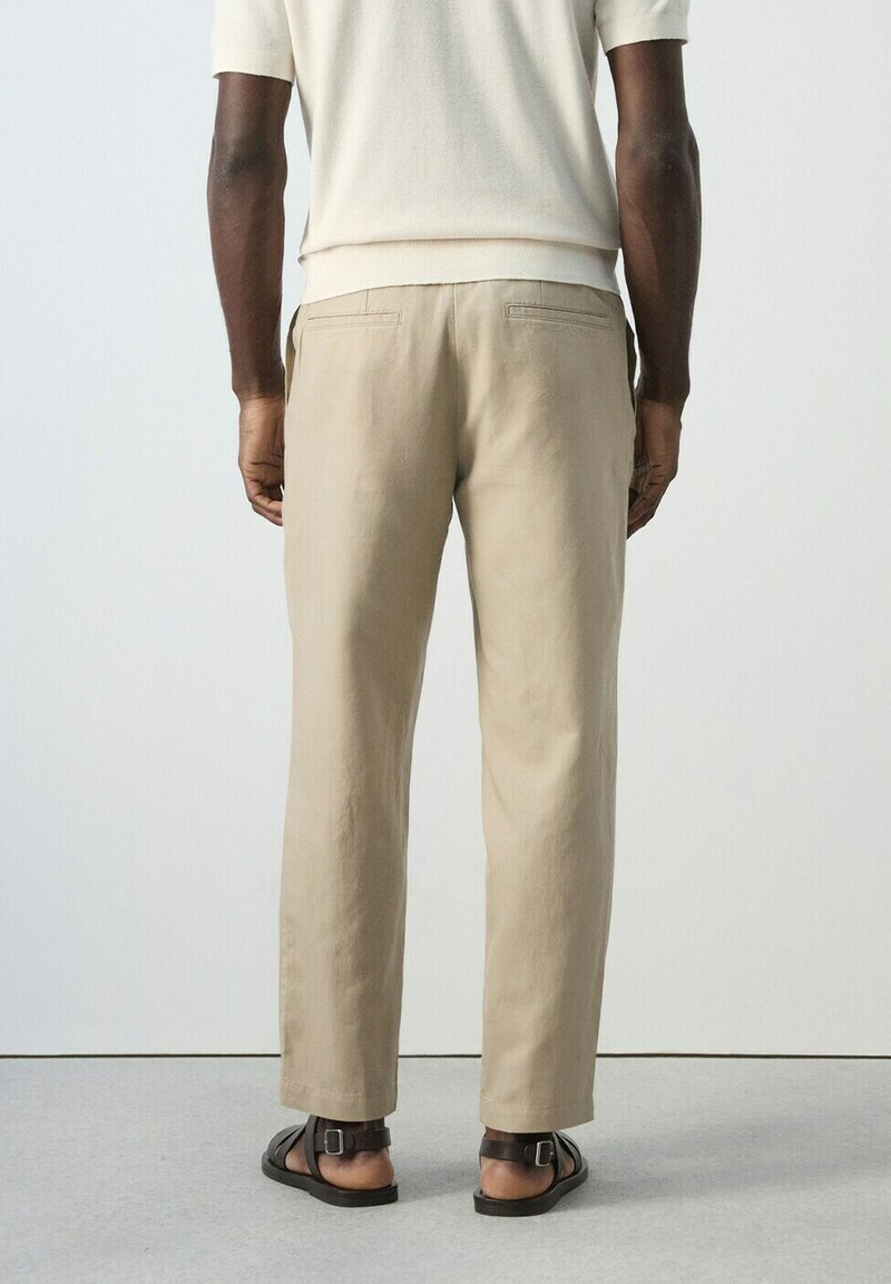 Man draagt beige broek, wit kortärmeligeshemd und dunkelbraune Sandalen, steht mit dem Rücken zu einer schlichten Kulisse.