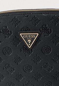 Μαύρη τσάντα από δέρμα με υφή, διαθέτει ανάγλυφα σχέδια και τριγωνική πλακέτα λογότυπου με την επιγραφή «GUESS U.S.A.» σε λευκό κείμενο και ένα ερωτηματικό.