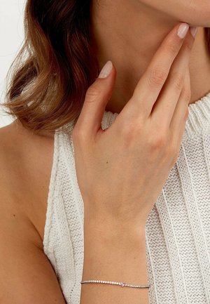 Woman's hand touching chin, wearing a delicate silver bracelet with a small pink stone, against white knitted sleeveless top background.