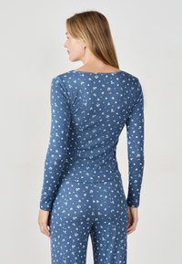 Kobieta z długimi blond włosami, ubrana w dopasowany niebieski, kwiatowy top z długimi rękawami i spodnie, zwrócona tyłem w stosunku do jednolitego tła.