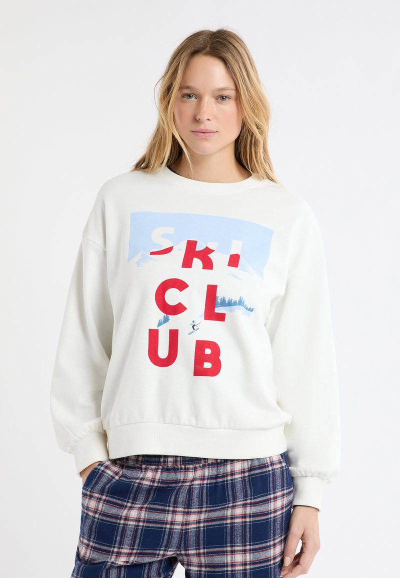 Biała bluza z grafiką "SKI CLUB" w kolorach czerwonym i niebieskim, z ilustracjami gór i narciarza. Mankiety i dół są wykończone ściągaczem.