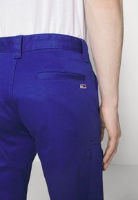 Modré bavlněné kalhoty s plochou zadní kapsou a malou nášivkou s logem. Mají rovný střih a hladkou texturu.