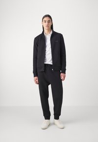 Mustmustu musta zip-up jakk, millel on seisev kaelus, kombineeritud jogger-pükstega ja valgete tossudega. Kangas näib sile ja sobib vabalt.