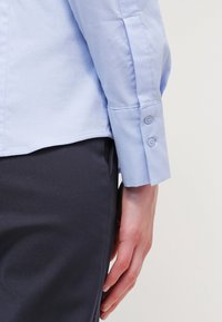 Camicia azzurro chiaro con colletto e polsini abbottonati, caratterizzata da una texture liscia e una vestibilità su misura, abbinata a pantaloni scuri.