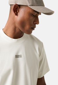 Lichtbeige katoenen T-shirt met een ronde hals, korte mouwen en een klein bruin logo op de borst. De stof lijkt glad te zijn.