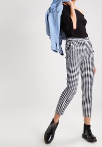 Schwarz-weiße Hosen im Pepita-Muster, knöchellang mit Taschen, kombiniert mit einem schwarzen, schulterfreien Oberteil und einer hellen Jeansjacke.