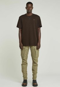 T-shirt in cotone marrone con maniche corte e collo rotondo, abbinato a pantaloni cargo verdi con molte tasche e una texture liscia.