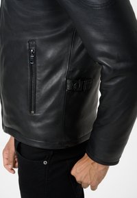 Schwarze Lederjacke mit einem seitlichen Reißverschlussfach, leichtem Glanz und glatter Textur. Inklusive eines gurtähnlichen Akzents an der Taille.