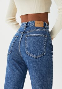 High-waisted blue denim jeans with a faded texture, featuring two back pockets and a rectangular PU leather label on the waistband.