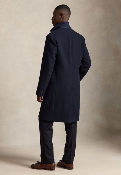 Homme de dos portant un long manteau bleu marine foncé, un pantalon sombre et des mocassins en cuir marron à glands, debout sur un fond neutre.