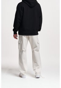 Personne debout de dos, portant un sweat à capuche noir, un pantalon cargo blanc avec poches latérales, et des baskets blanches sur un fond blanc uni.