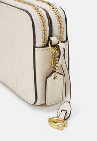 Kremowa skórzana saszetka na zamek z złotymi elementami, kontrastującą brązową lamówką, fakturowaną powierzchnią i ozdobną zawieszką przymocowaną łańcuszkiem z koralikami.