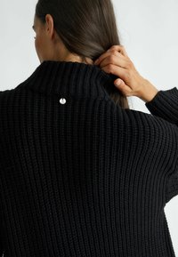 Černý žebrovaný pletený svetr s vysokým límcem. Vzadu je malý stříbrný dekorativní knoflík. Texturovaný materiál s volným střihem.