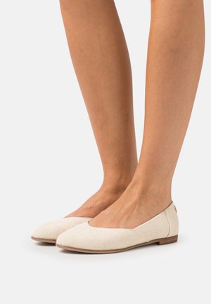 Jambes portant des ballerines en tissu texturé couleur crème avec des talons en bois bas sur un fond blanc.