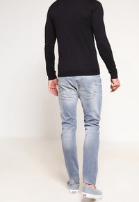 Camisa negra de manga larga con diseño ajustado, combinada con jeans ajustados de color azul claro que presentan un sutil desgaste y dos bolsillos traseros. Zapatos grises.