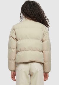 Veste rembourrée beige avec un col montant et des poignets élastiques. Présente des coutures horizontales et une silhouette cropped. Texture lisse et brillante.