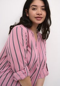 Rosa randig blus gjord av lätt tyg. Har grå vertikala ränder, en knappslå och uppkavlade ärmar.