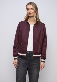 Femme portant une veste bomber bordeaux avec des poignets rayés blancs, une chemise blanche et un jean gris foncé, debout devant un fond uni gris clair.