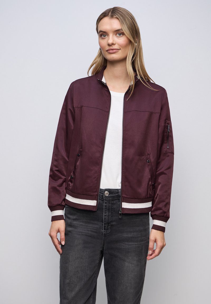 Femme portant une veste bomber bordeaux avec des poignets rayés blancs, une chemise blanche et un jean gris foncé, debout devant un fond uni gris clair.