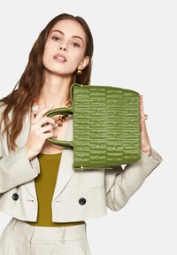 Grön texturerad handväska med upphöjt mönster, kontrasterande handtag och guldaccenter, hålls av en modell som bär en ljus kavaj och en olivfärgad topp.