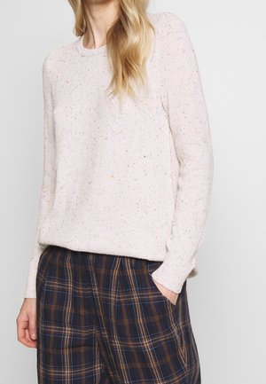 Femme portant un pull en maille clair moucheté associé à un pantalon à carreaux foncé, une main dans la poche, sur un fond uni.