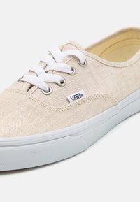 Baskets en toile beige clair avec des lacets blancs, des œillets en métal et une texture à motifs. Elles comportent une semelle en caoutchouc blanche et le logo Vans.