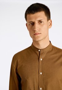 Camisa marrón de botones con cuello alto, fila única de botones blancos, textura suave y mangas largas.