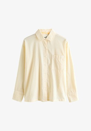 Woman adjusting loose-fitting pale yellow striped pajamas standing beside a bed with blue and pink striped bedding.