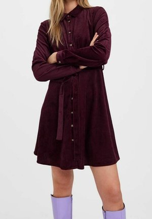 Femme portant une robe en velours côtelé bordeaux à manches longues boutonnée avec une ceinture à la taille, associée à des bottes lavande montant jusqu'aux genoux, les bras croisés.