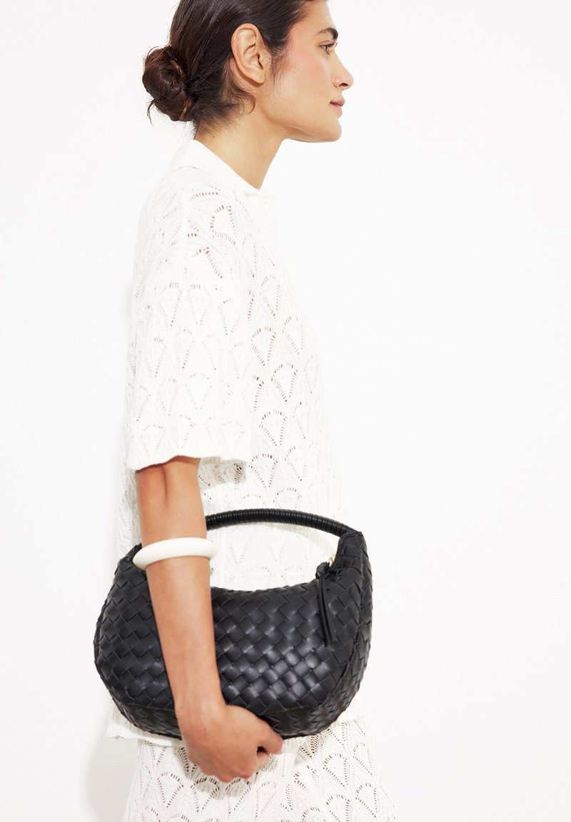 Svart flätad handväska med rund form och kort handtag, med en texturerad design och dragkedjestängning, hålls av en person som bär en vit topp.